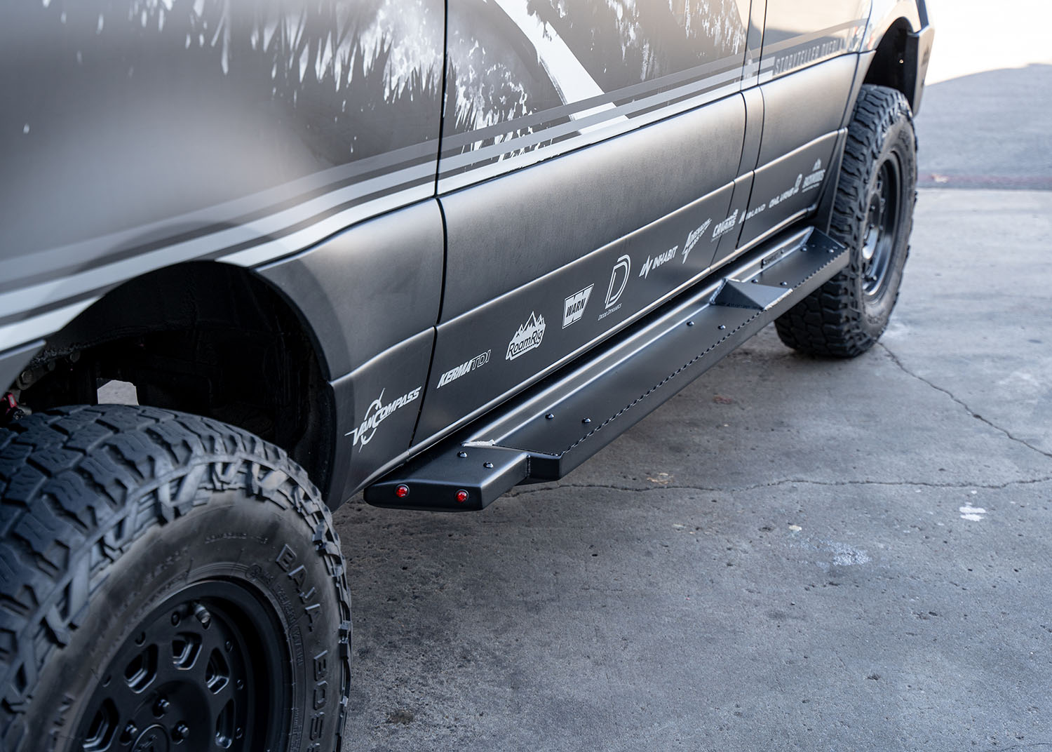 Side view of ROAMBUILT Flat Side Step in black textured aluminum, mounted on a Mercedes Sprinter, showing raised grip edge and lights.
