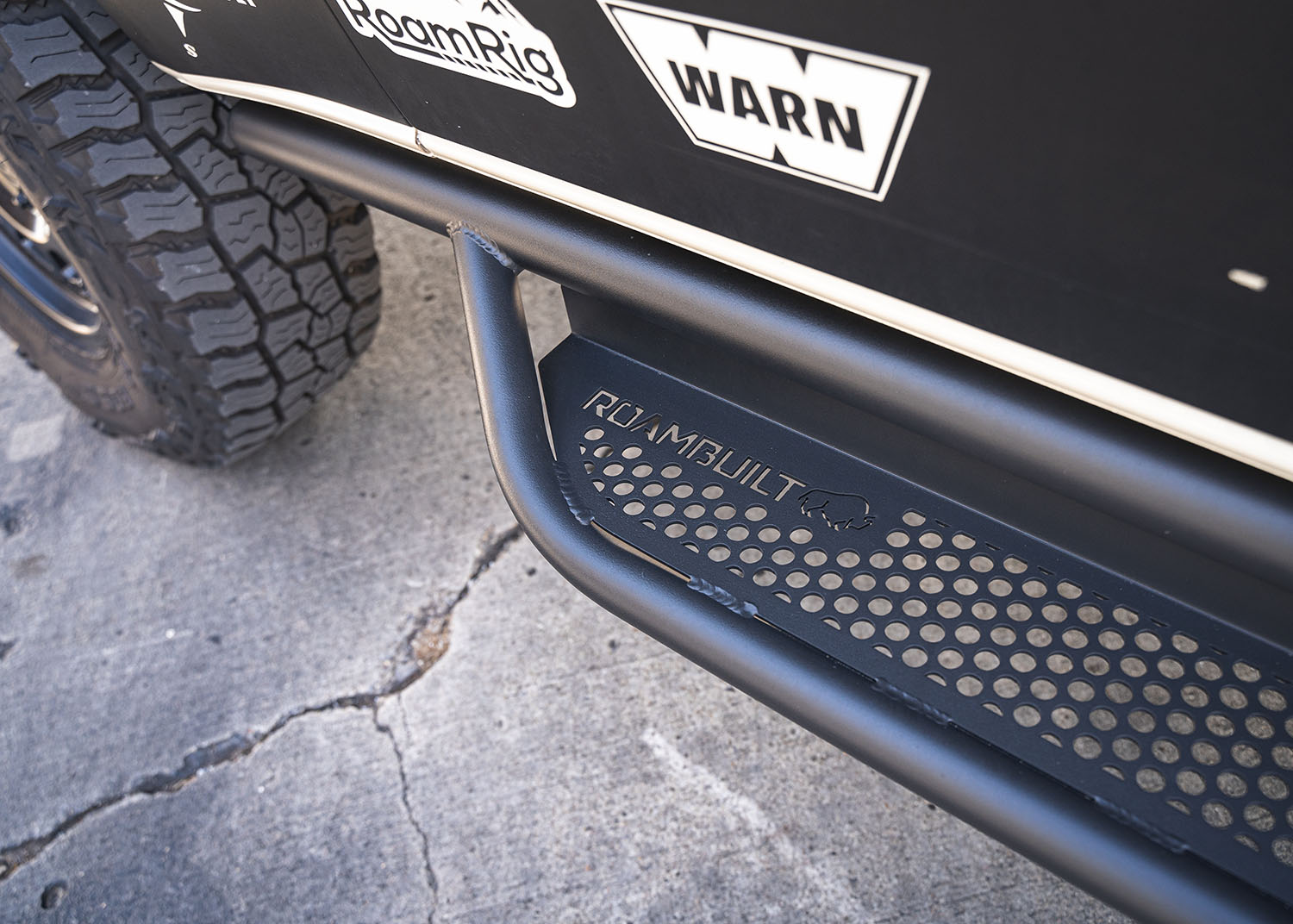 Close-up view looking down at the rear section of the ROAMBUILT Passenger Side Tube Step, highlighting the logo and perforated tread surface.