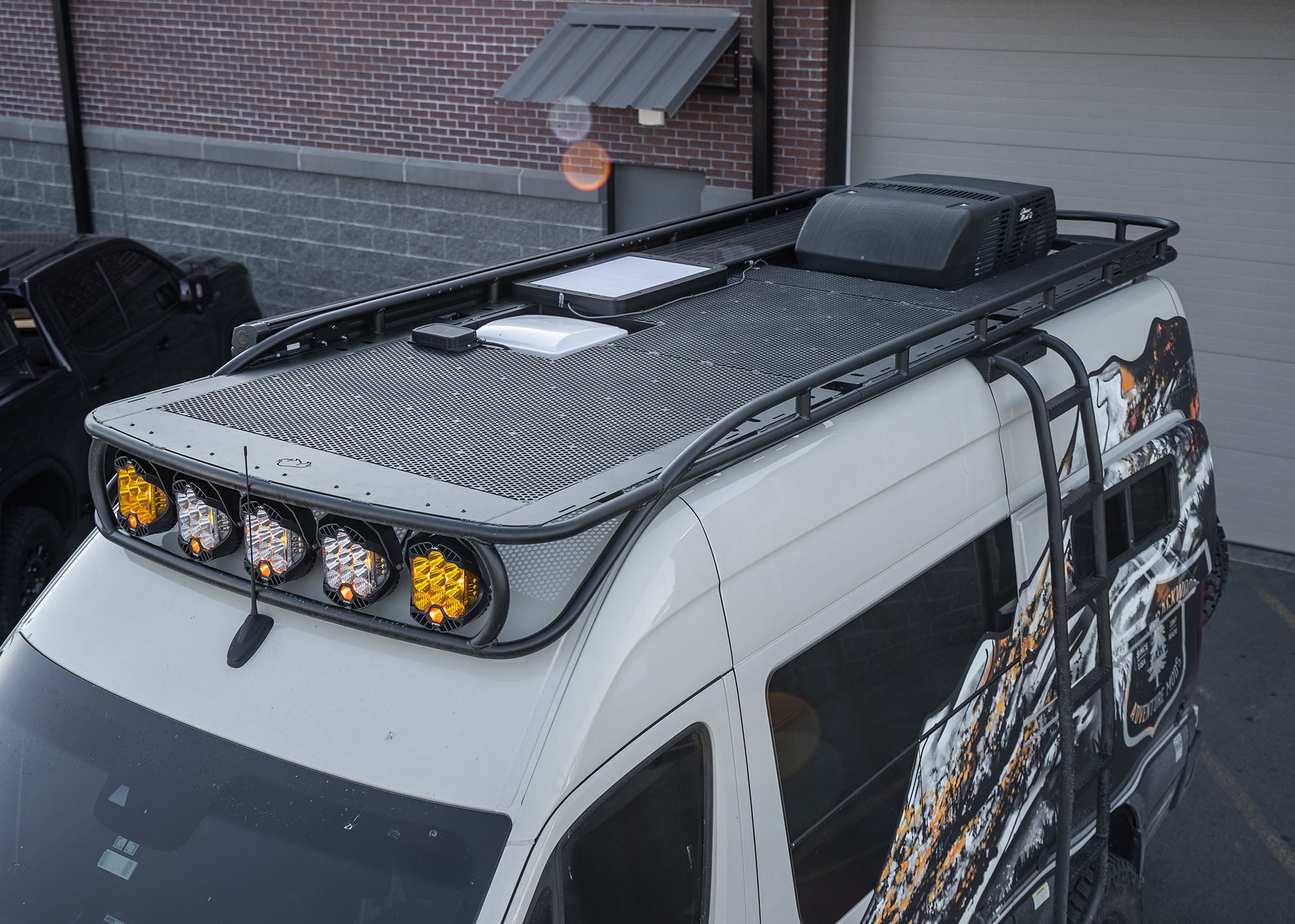 Front and top driver-side view of the Safari 2.0 Roof Rack mounted on a 2007+ Mercedes Sprinter, showing curved front profile and perforated aluminum decking.