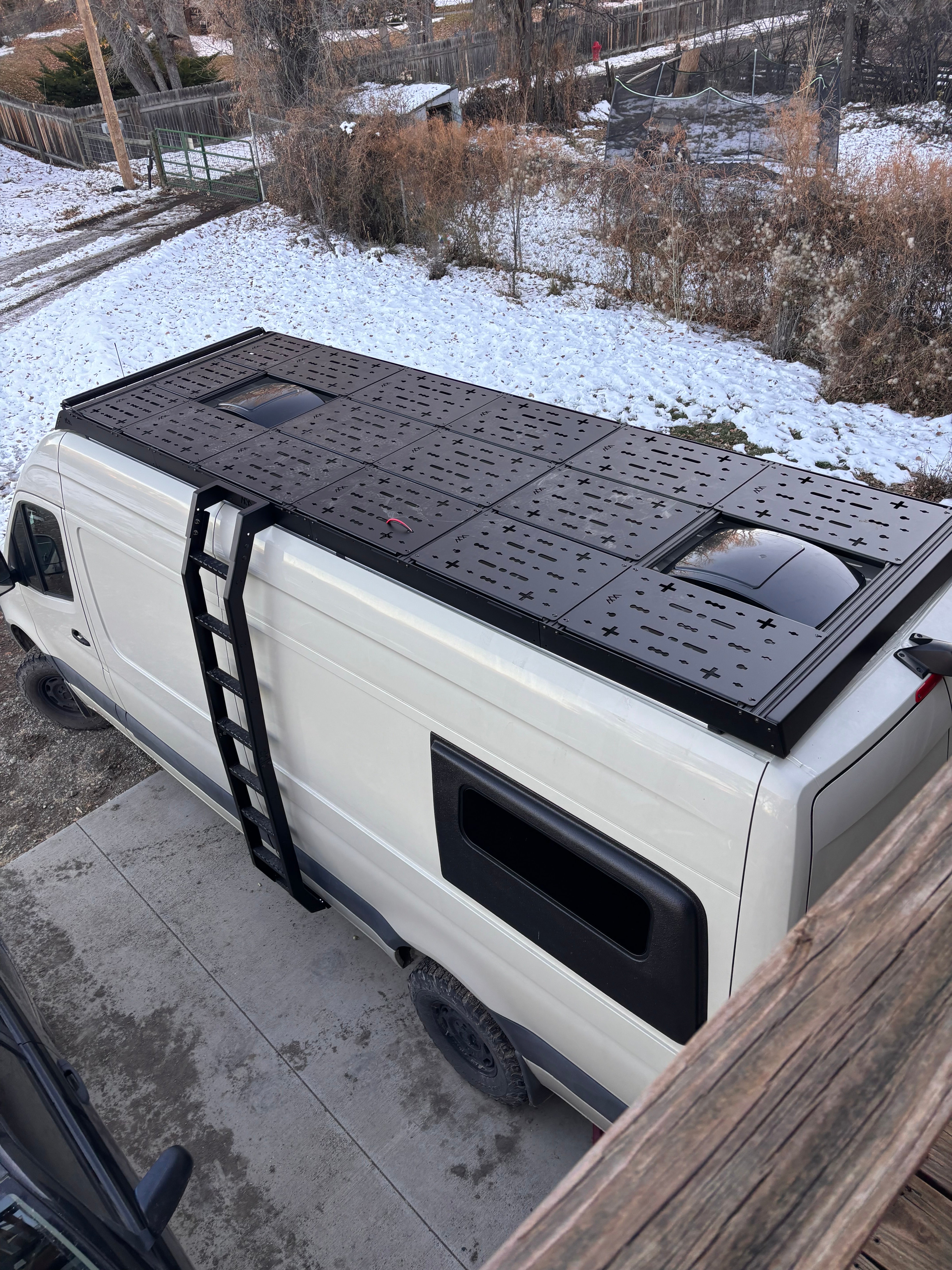 Driver-side rear drone view of van roof with ROAMBUILT deck panels installed, showing rooftop storage setup and lounging area configuration.