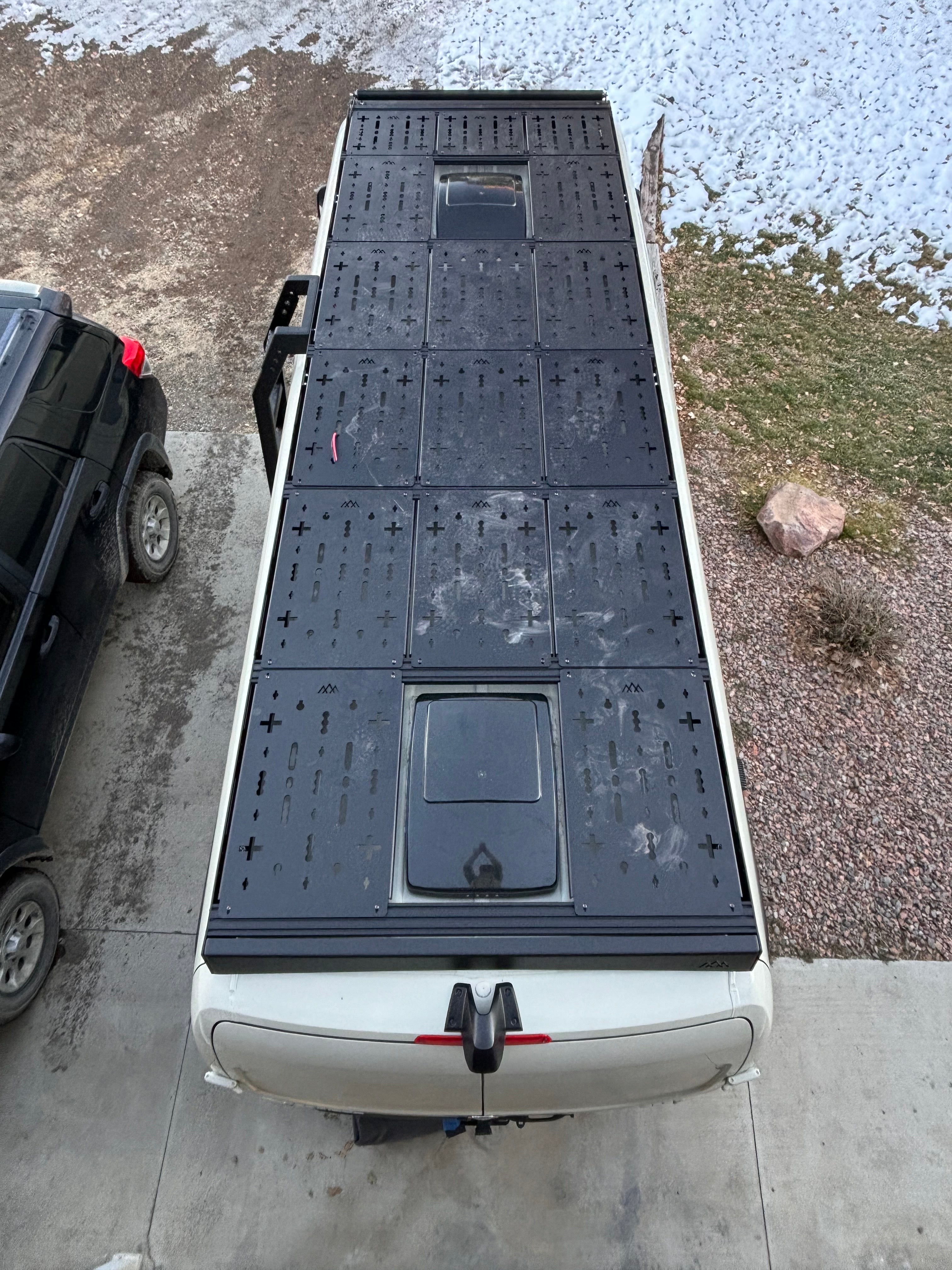 Rear drone view of entire van roof outfitted with ROAMBUILT deck panels, showing full layout from back to front for storage and rooftop lounging.