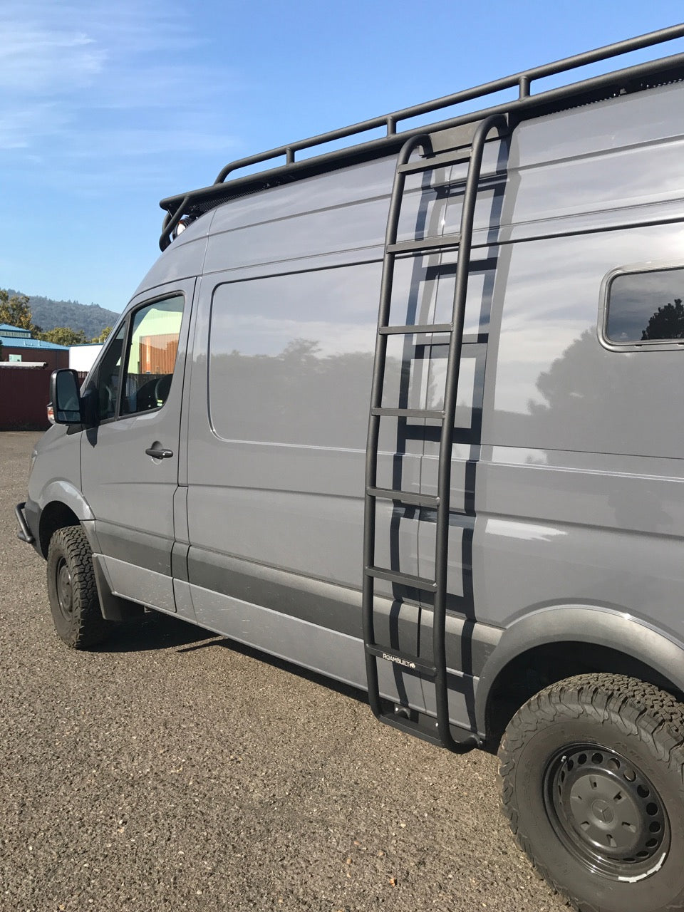 Driver side view of a Mercedes Sprinter van with the full ROAMBUILT Straight Side Ladder mounted, showing the complete ladder length, mounting brackets, and integration with the vehicle’s roof rail and body.